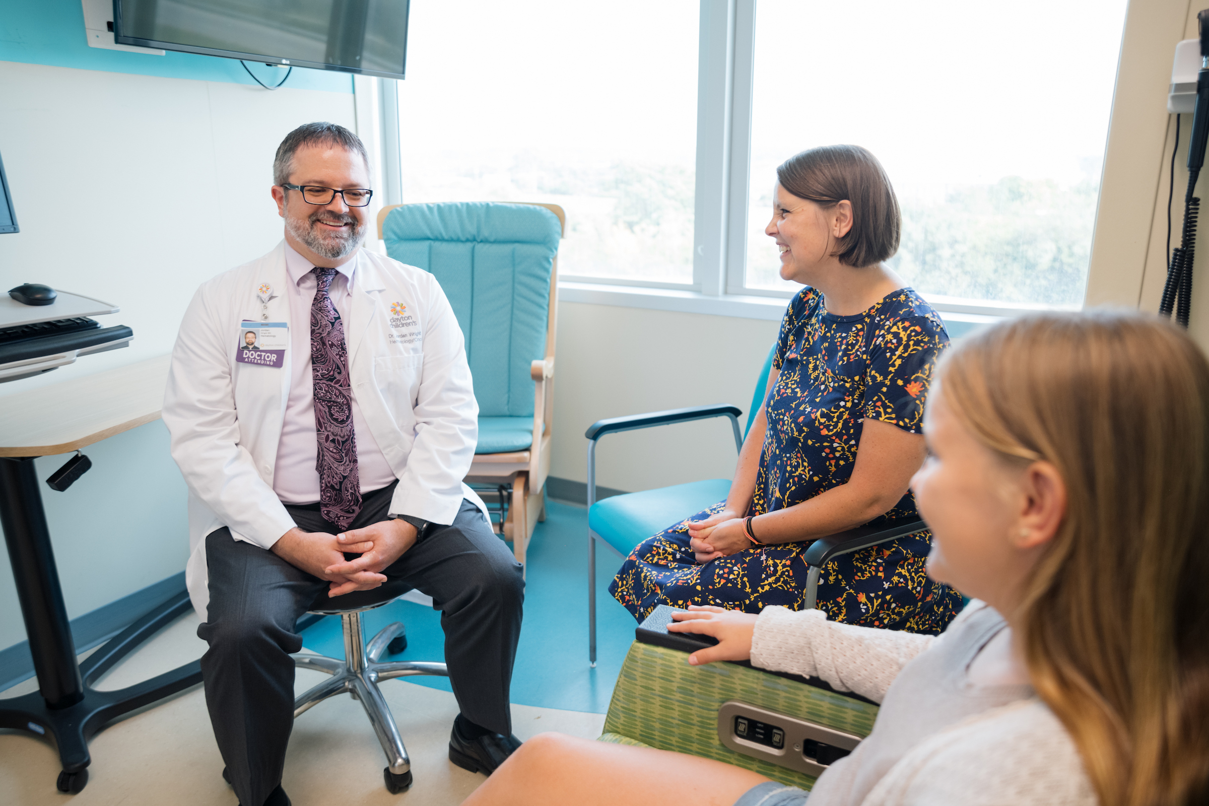 Doctor meets with mom and young girl for a follow-up oncology visit in bright exam room.