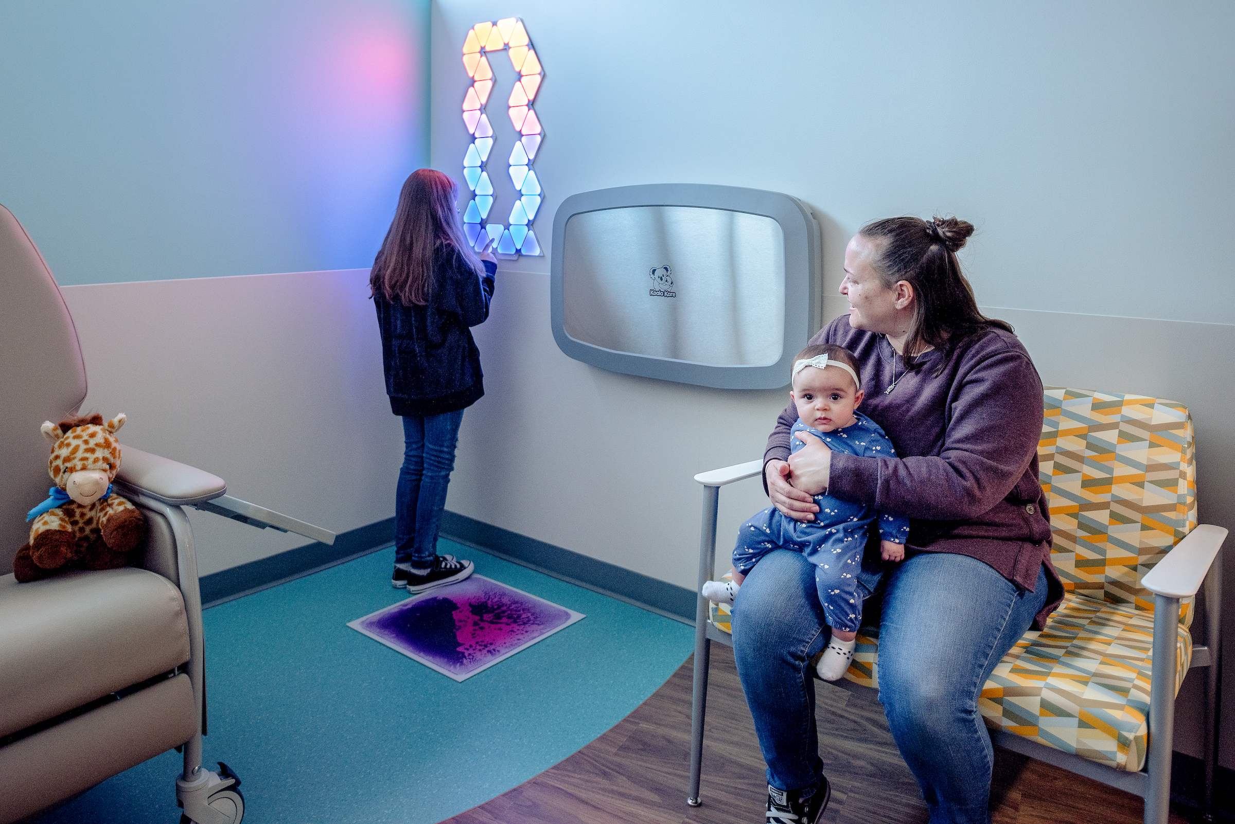 Young patient playing with sensory-friendly light on wall with mom and baby sibling before lab draw.