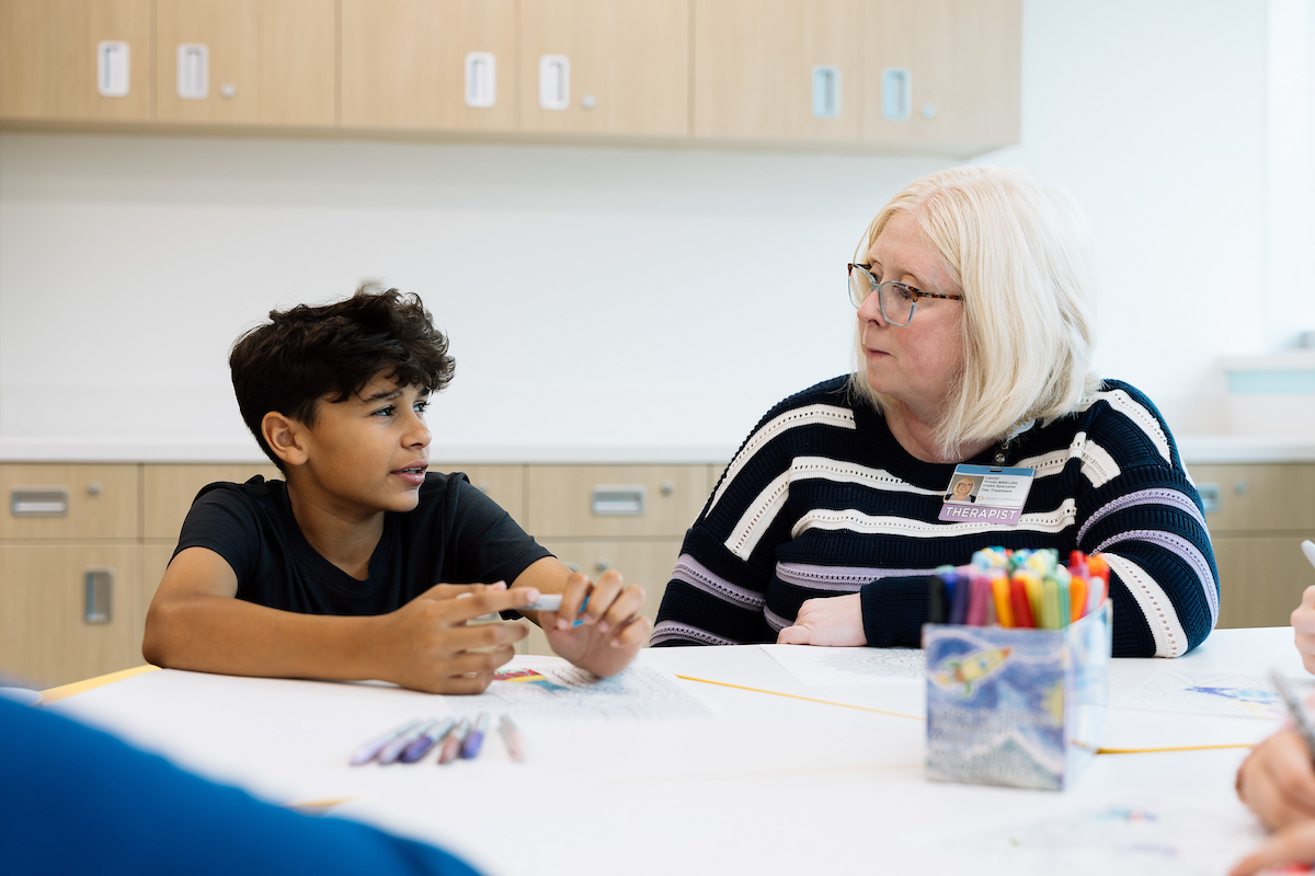 Female therapist speaking with teen boy at a table together while he draws.