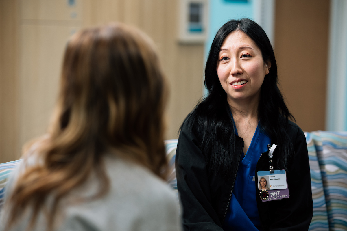 Mental health technician sits with teen on a couch talking together.